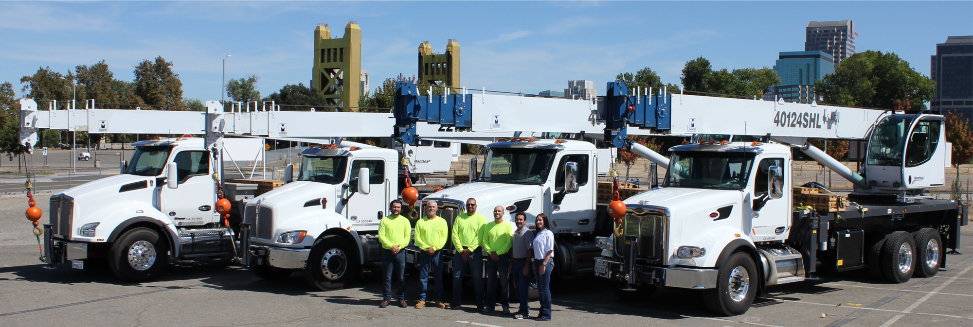 Three large utility trucks from a local crane rental company line up in a parking area. Six people, wearing safety gear, stand in front of the vehicles. In the background, there’s a city skyline and a distinctive yellow bridge.