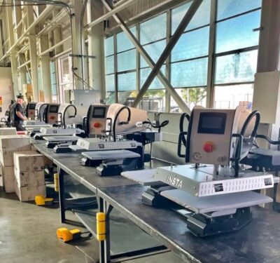 Row of Insta Graphic Systems heat press machines set up in a bright industrial workspace with large windows and organized workstations