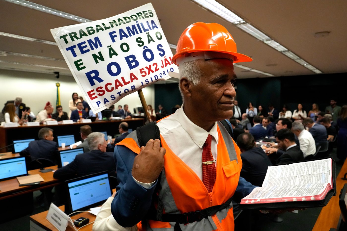 Lamaker and Pastor Sargento Isidorio holds a sign supporting the end of Brazil's six-day workweek schedule, that reads in Portuguese: "Workers have families and are not robots. 5x2 schedule now," during a special committee session analyzing the proposal at the Chamber of Deputies in Brasilia, Brazil, Wednesday, May 27, 2026. (AP Photo/Eraldo Peres)