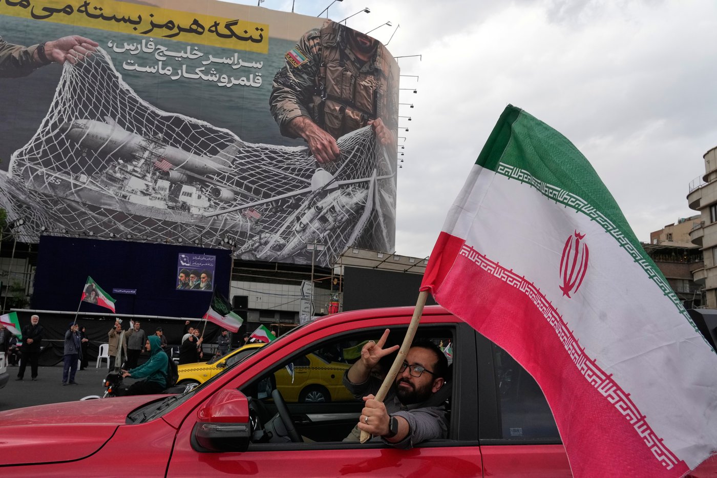 A man flashes a victory sign as he carries an Iranian flag in front of an anti-U.S. billboard depicting the American aircrafts into the Iranian armed forces fishing net with signs that read in Farsi: "The Strait of Hormuz will remain closed, The entire Persian Gulf is our hunting ground," at the Eqelab-e-Eslami, or Islamic Revolution Square in downtown Tehran, Iran, Monday, April 13, 2026. (AP Photo/Vahid Salemi)