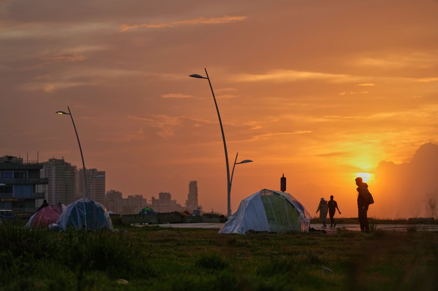 People walk past tents sheltering people displaced by Israeli airstrikes at a public space along the Beirut waterfront at sunset in Beirut, Lebanon, Sunday, March 15, 2026. (AP Photo/Hassan Ammar)