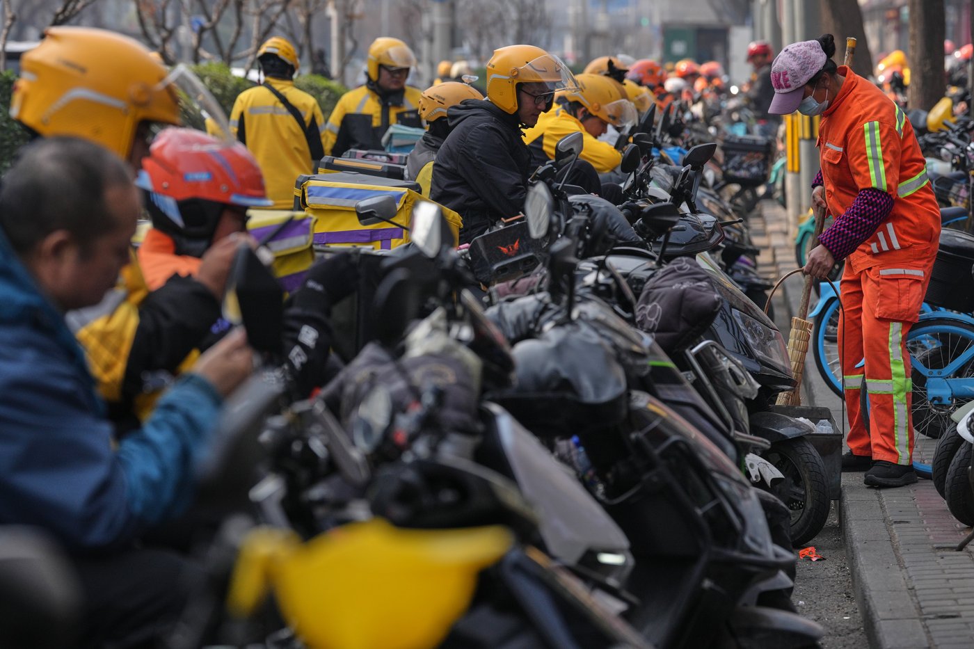 A cleaner sweeps near food delivery riders gather outside restaurants waiting for online orders, in Beijing, China, Wednesday, March 25, 2026. (AP Photo/Andy Wong)