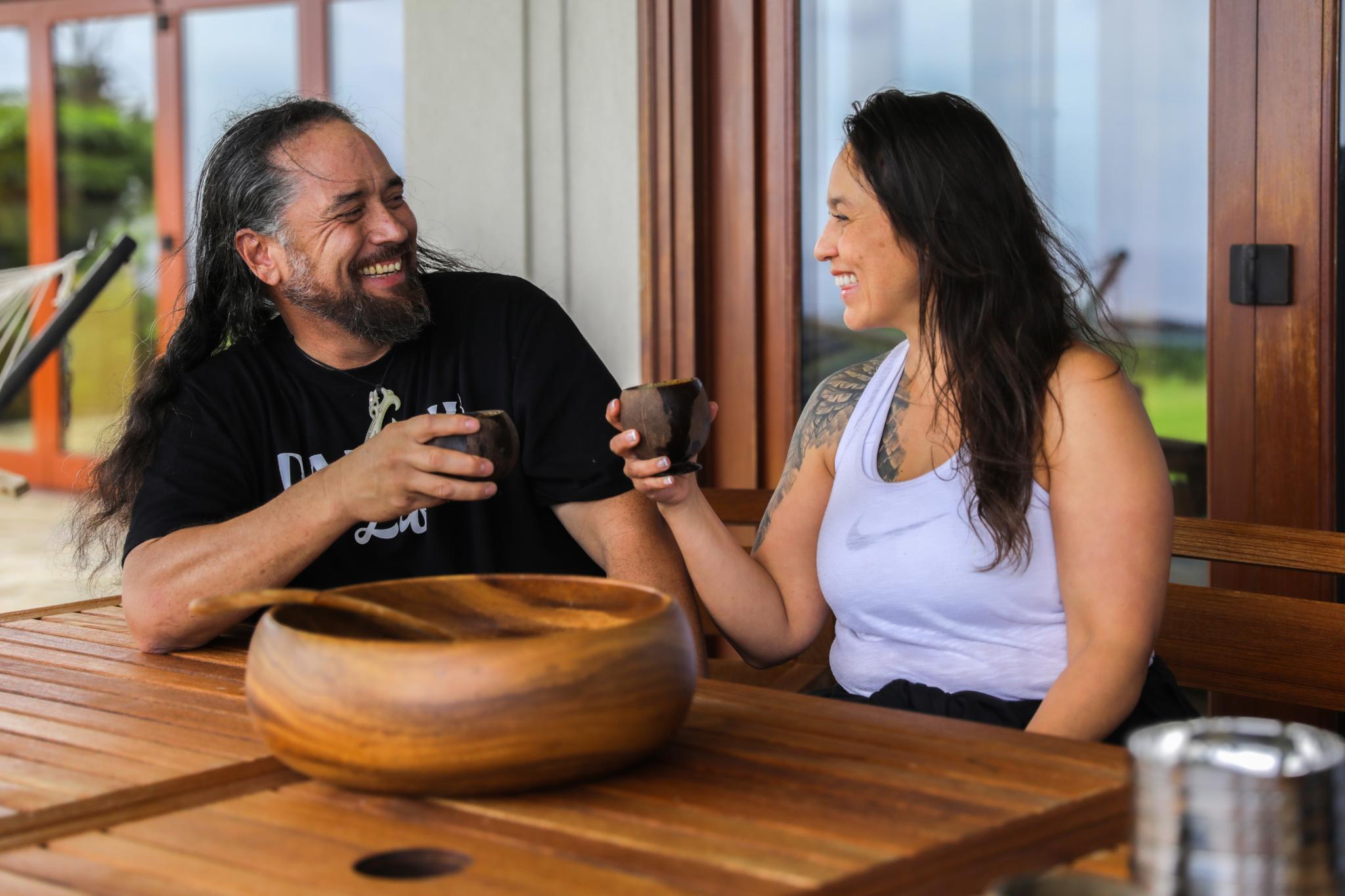 Two adults sitting at a wooden table smiling and clinking small cups of kava with a large wooden bowl in front of them.