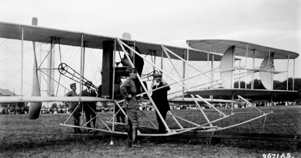 Charlie Taylor pours fuel into the gas tank of the Flyer while Orville Wright goes over the proposed flight route with his passenger, Lt. Frank Lahm. (Photo: wright-brothers.org)