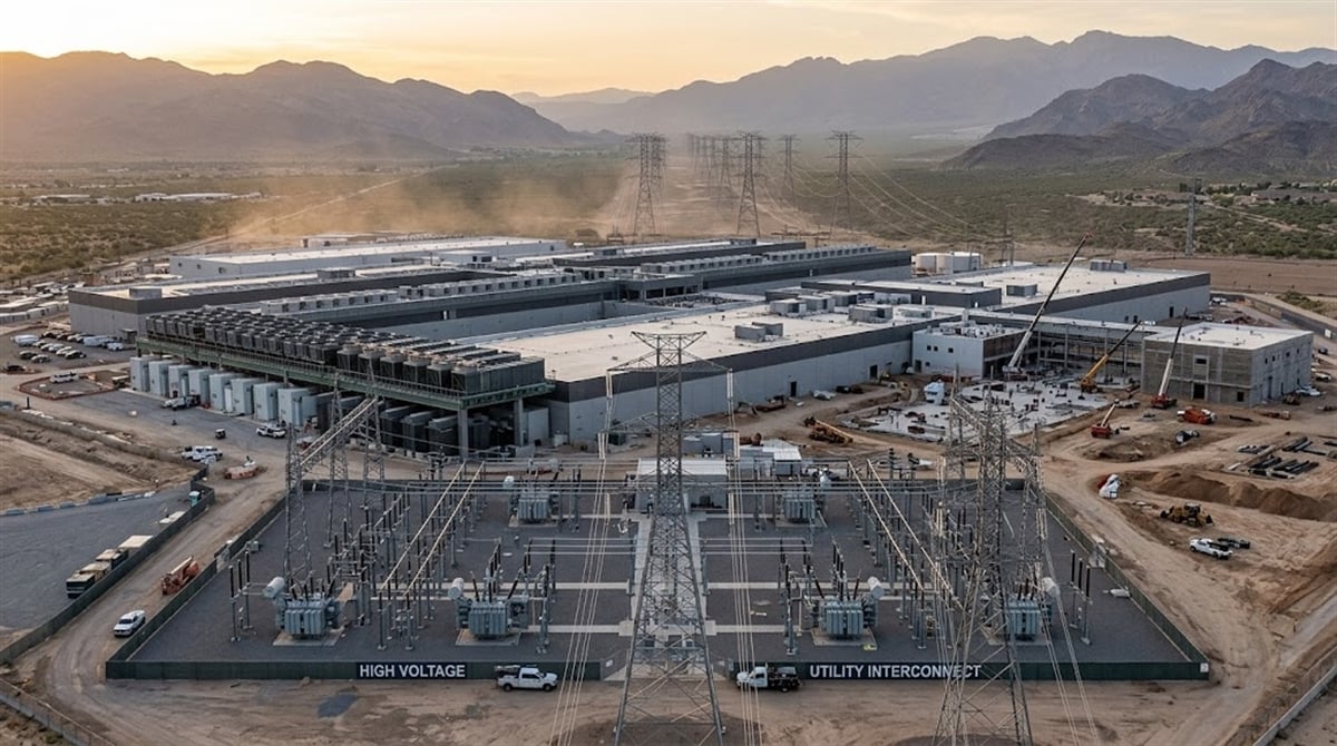 Aerial view of a large data center campus under construction in a desert setting with a high-voltage utility substation in the foreground.