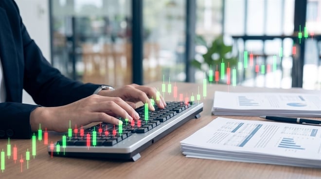 A person in business attire types on a keyboard beside financial reports with overlaid candlestick chart graphics.