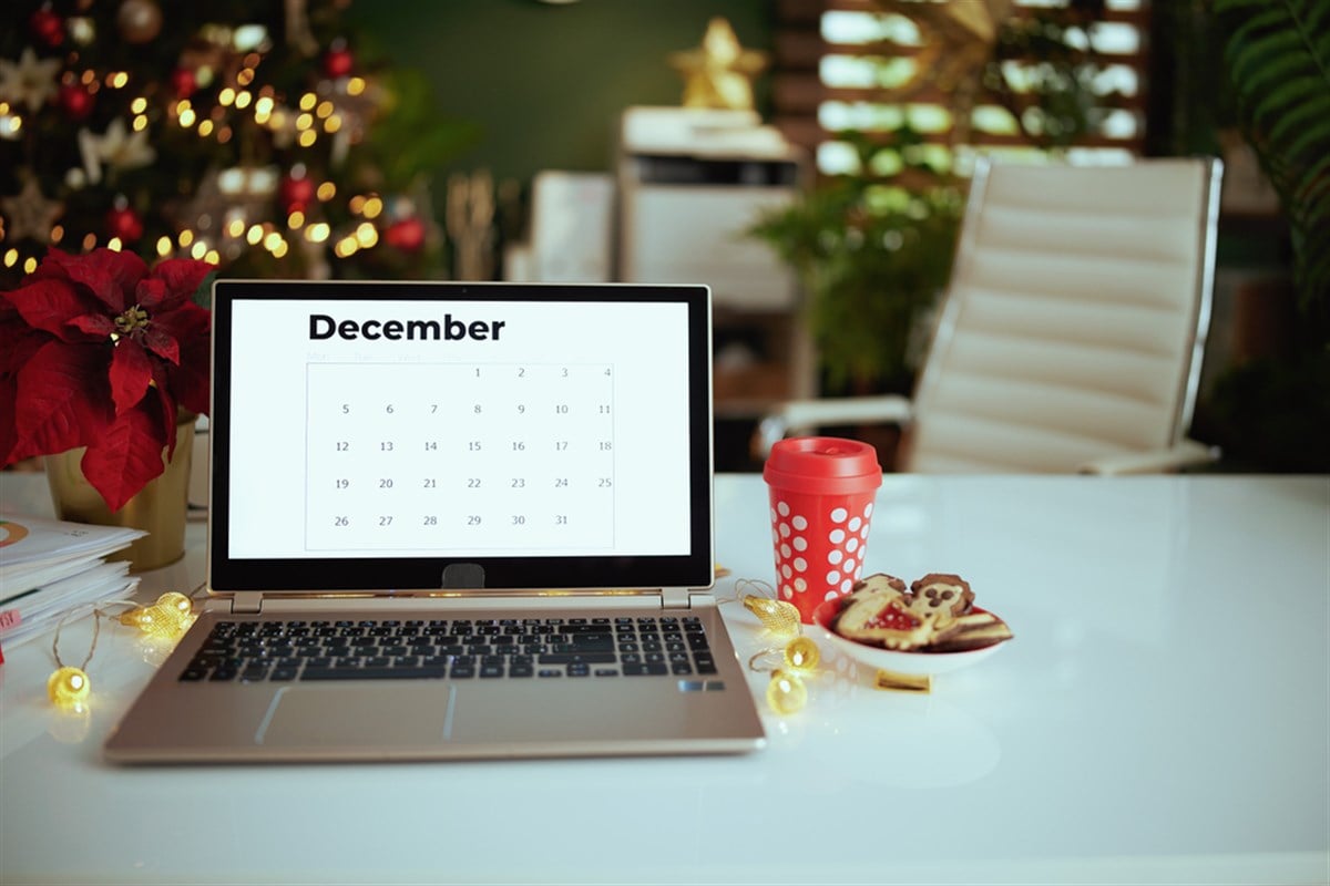 Office with December calendar on laptop screen, poinsettia, Christmas beverage and cookies.