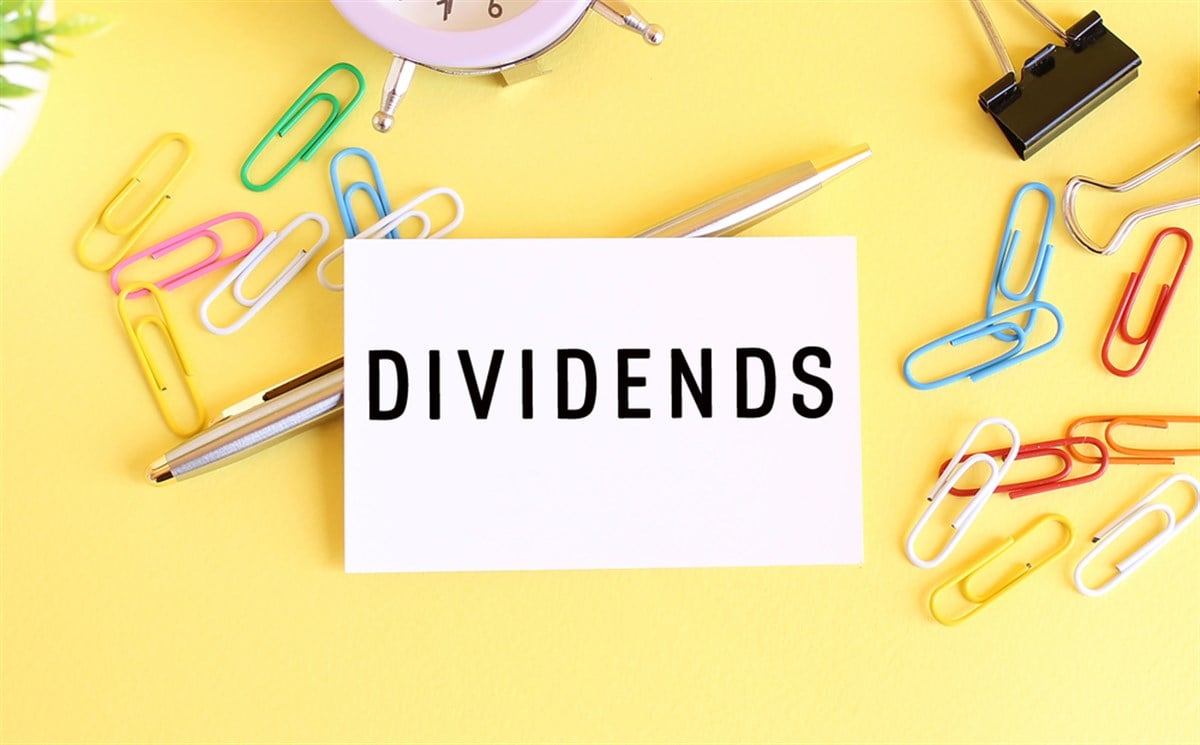 Top view of a business card with text DIVIDENDS, pen, paper clips and clock on a yellow background. Business concept. — Photo