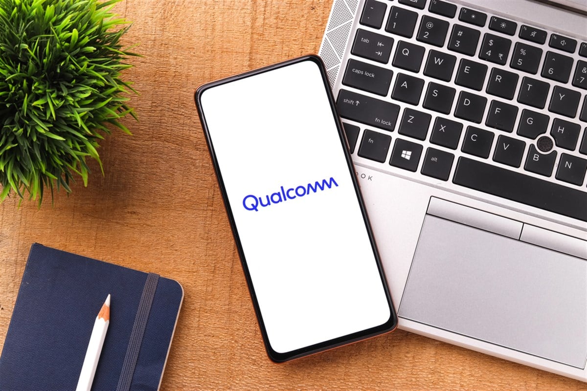 Photo of qualcomm logo on phone screen, phone sitting on a desk next to a laptop, notebook, pencil
