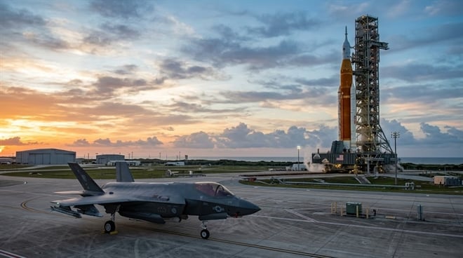 An F-35 fighter jet sits on a tarmac near NASA's Space Launch System rocket on a launch pad at sunset.