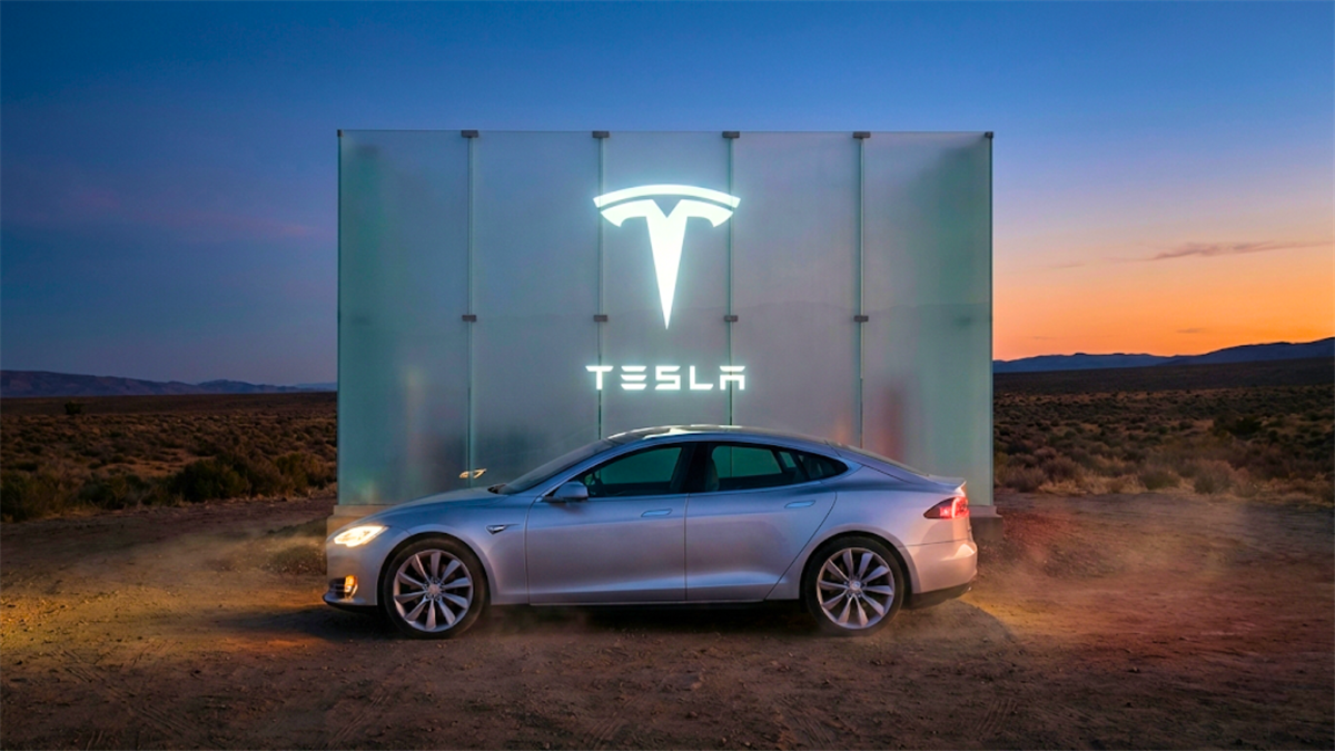 Silver Tesla sedan parked in a desert at dusk with a large glowing Tesla logo behind it.