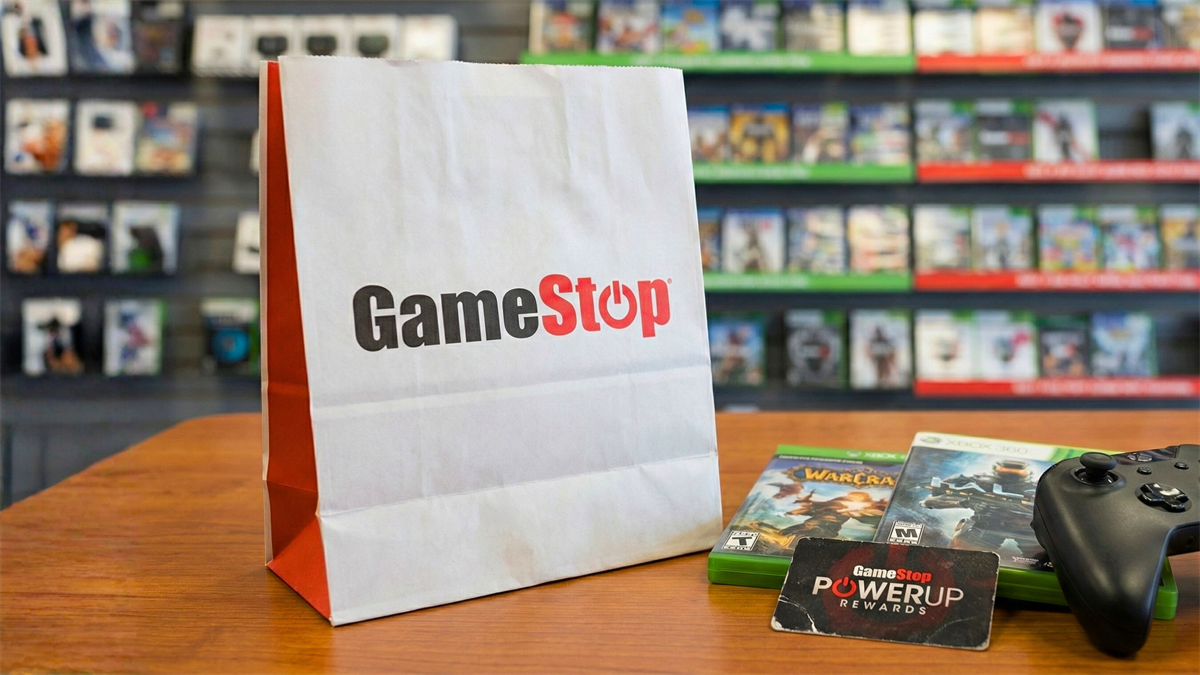 A GameStop bag on a counter with games, a rewards card, and a controller in front of blurred store shelves.