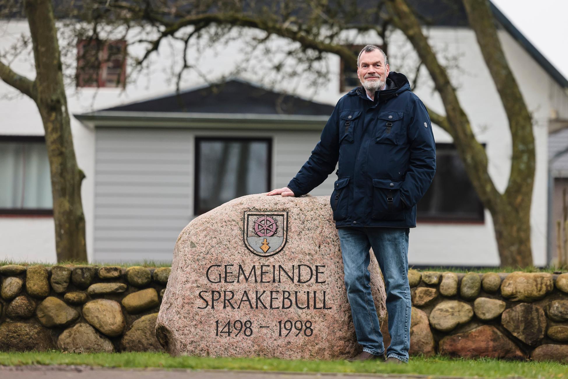 Juergen Hansen, Mayor of the village of Sprakebuell, stands next to a stone with the name of the municipality in Sprakebuell, Germany, Thursday, March 14, 2024. Sprakebuell is something of a model village for the energy transition - with an above-average number of electric cars, a community wind farm and renewable heat from biogas. All houses in the village center have been connected to the local heating network and all old oil heating systems have been removed. (AP Photo/Frank Molter)