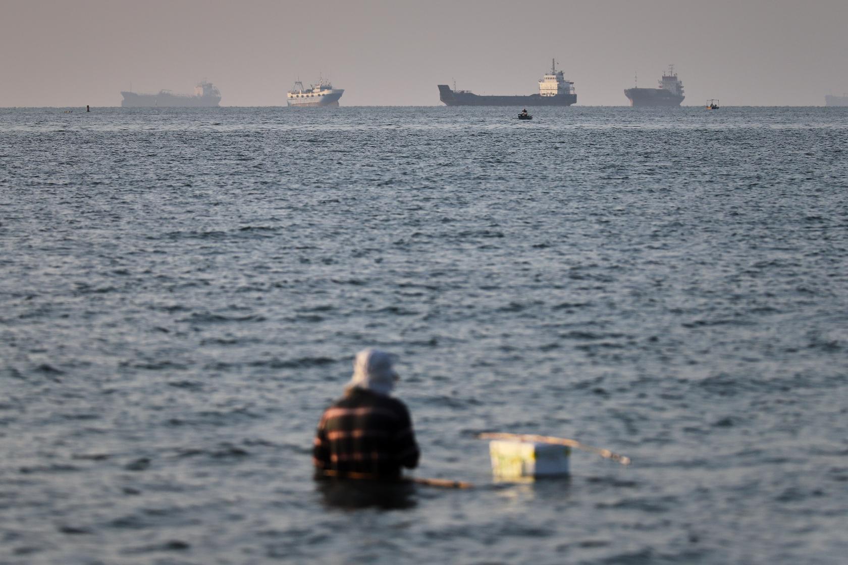 A person sits in shallow water as cargo and commercial vessels are anchored in the Strait of Hormuz off Bandar Abbas, Iran, Monday, June 8, 2026. (Amirhosein Khorgooi/ISNA via AP)