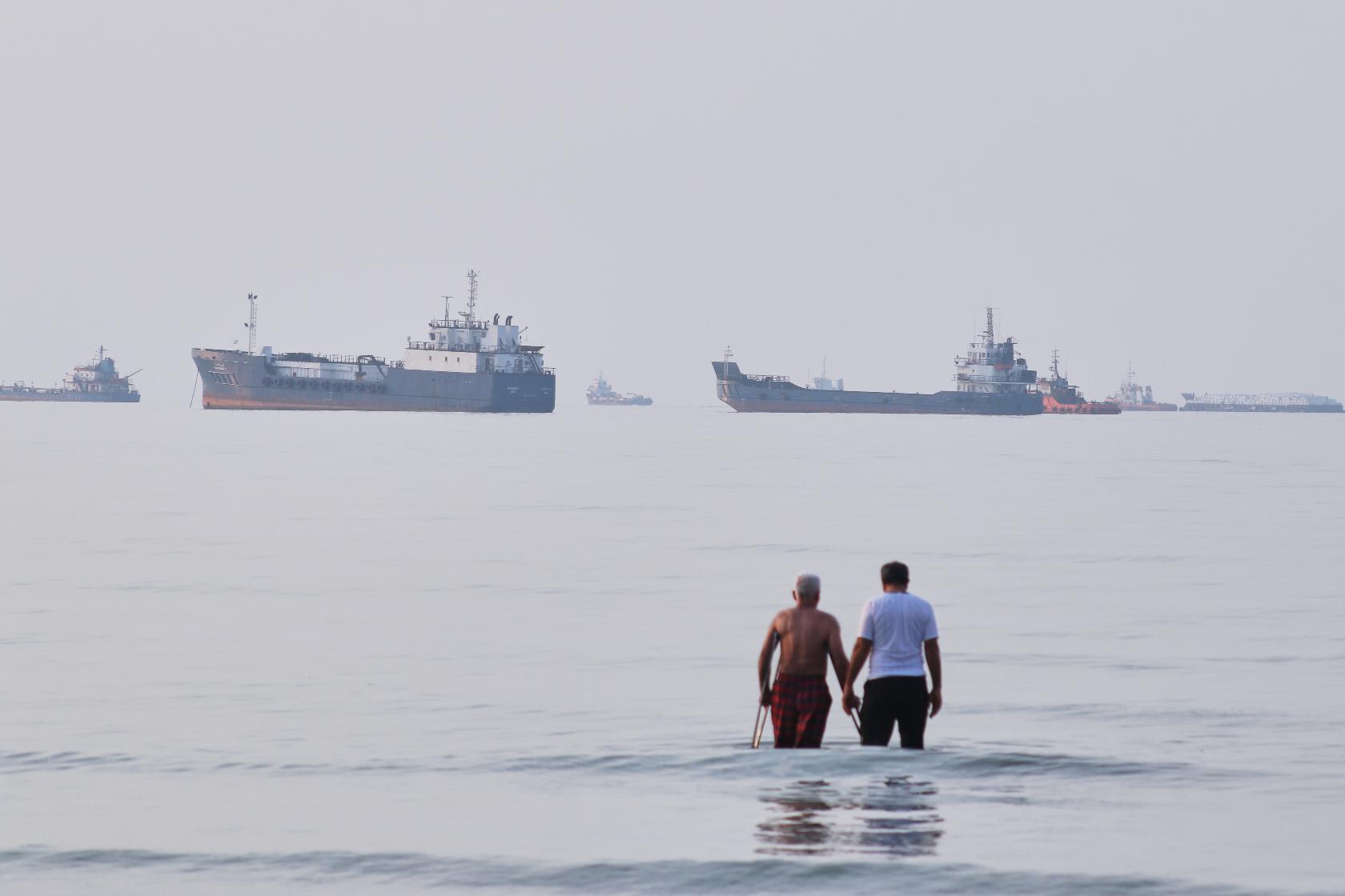 Two men wade in the waters of the Strait of Hormuz with vessels anchored in the background, off Bandar Abbas, Iran, Sunday, July 12, 2026. (Razieh Poudat/ISNA via AP)