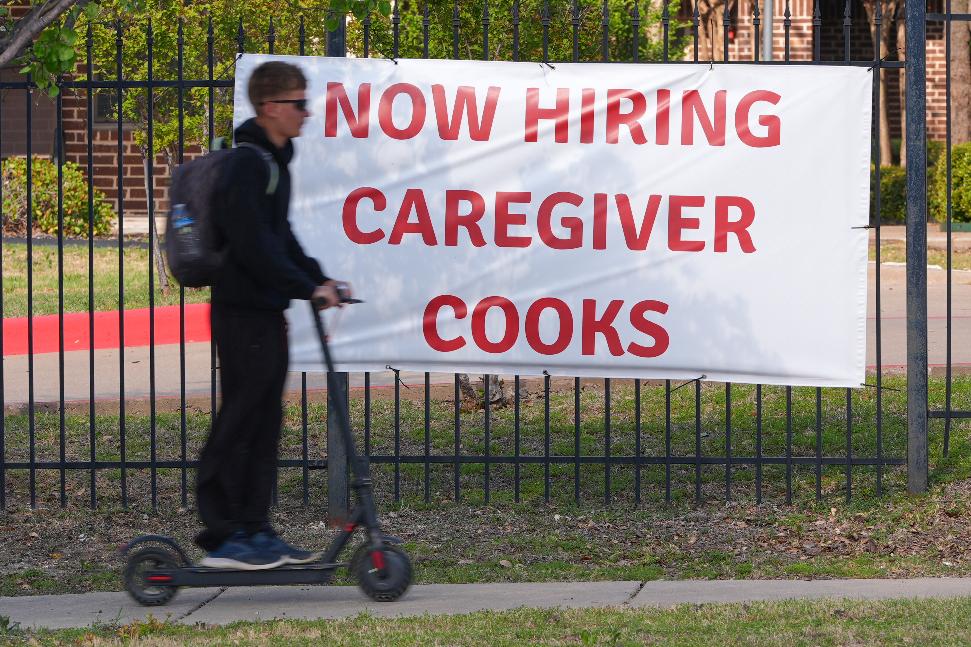 A now hiring sign sits by the sidewalk as a rider on a scooter passes in Garland, Texas, Monday, March 23, 2026. (AP Photo/LM Otero)