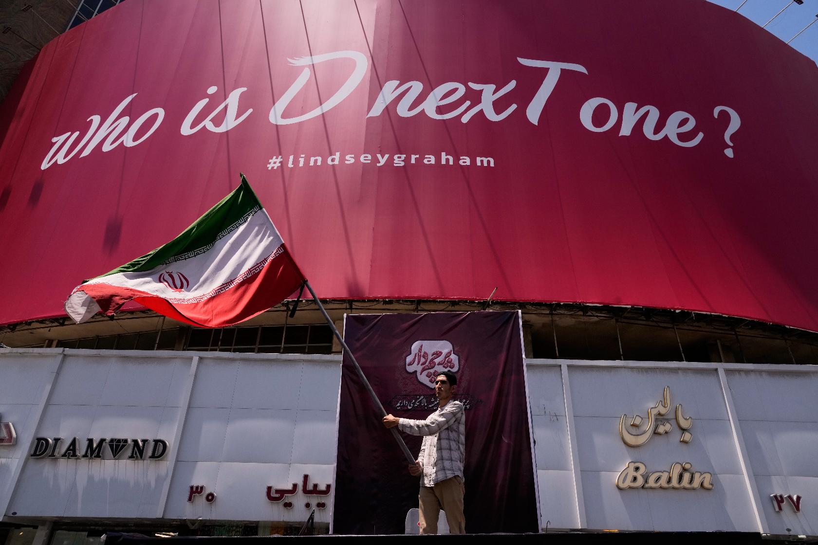 A man waves an Iranian flag beneath a billboard reading in English, "Who is D nexT one?" and "#lindseygraham," referring to late U.S. Sen. Lindsey Graham and using the capital letters "D" and "T" in an apparent play on the initials of U.S. President Donald Trump, in downtown Tehran, Iran, Thursday, July 16, 2026. (AP Photo/Vahid Salemi)