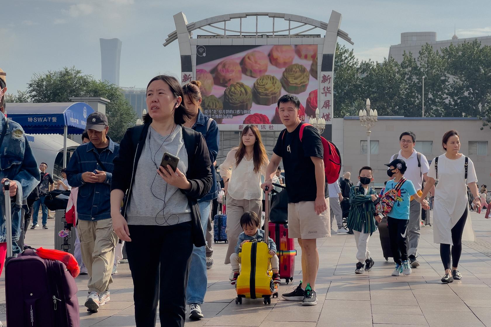 Travelers walk by a screen showing the mooncakes as they arrive at Beijing Railway Station to catch their trains in Beijing, Friday, Sept. 29, 2023. Tens of millions of Chinese tourists are expected to travel within their country, splurging on hotels, tours, attractions and meals in a boost to the economy during the 8-day autumn holiday period that began Friday. (AP Photo/Andy Wong)