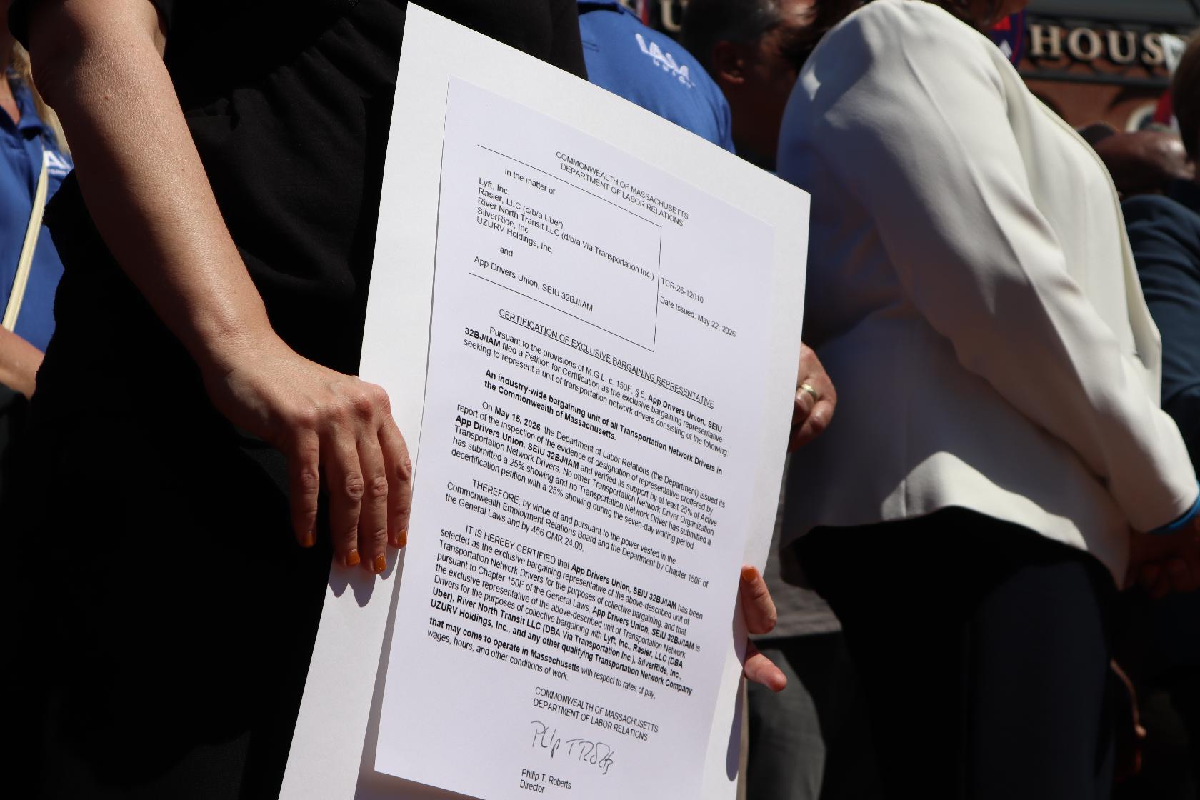 A demonstrator holds copy of a certification recognizing the App Drivers Union as the bargaining representative for Massachusetts rideshare drivers during a rally outside the Massachusetts State House after the App Drivers Union announced it had become the first certified union of rideshare drivers in the nation, on Tuesday, May 26, 2026, in Boston. (AP Photo/Leah Willingham)