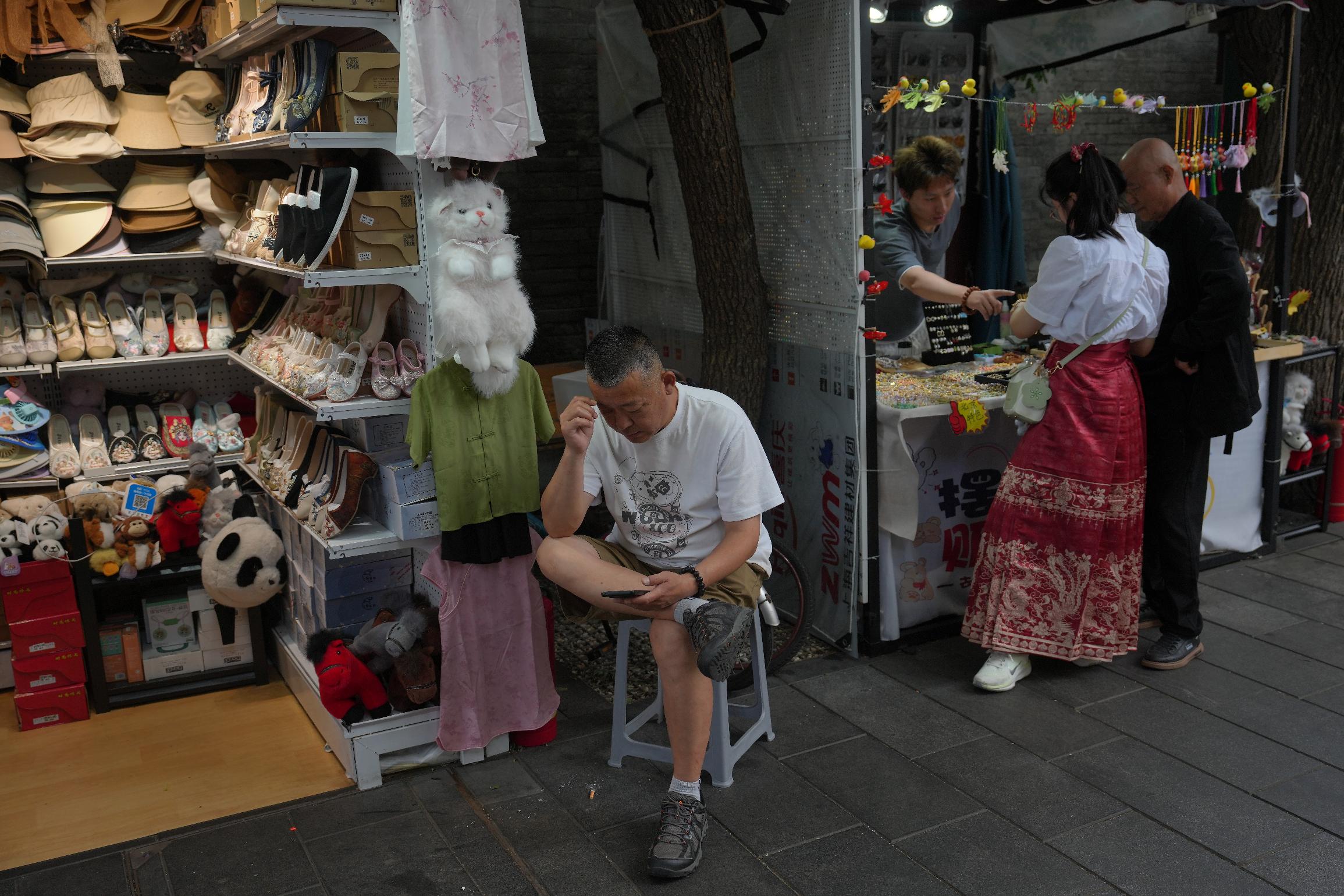 A vendor browses a smartphone as another attends customers at their stores selling souvenirs at Nanluoguxiang, a popular tourist spot in Beijing, Wednesday, June 24, 2026. (AP Photo/Andy Wong)